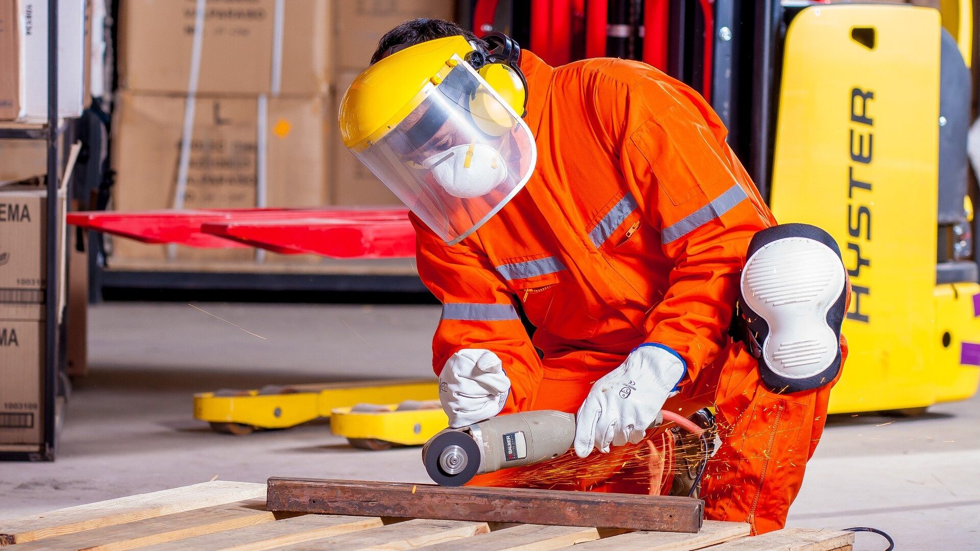 Operaio inginocchiato a tagliare con una fresa una barra di metallo su pallet di legno con addosso tuta da lavoro arancione catarifrangente e occhiali protettivi.