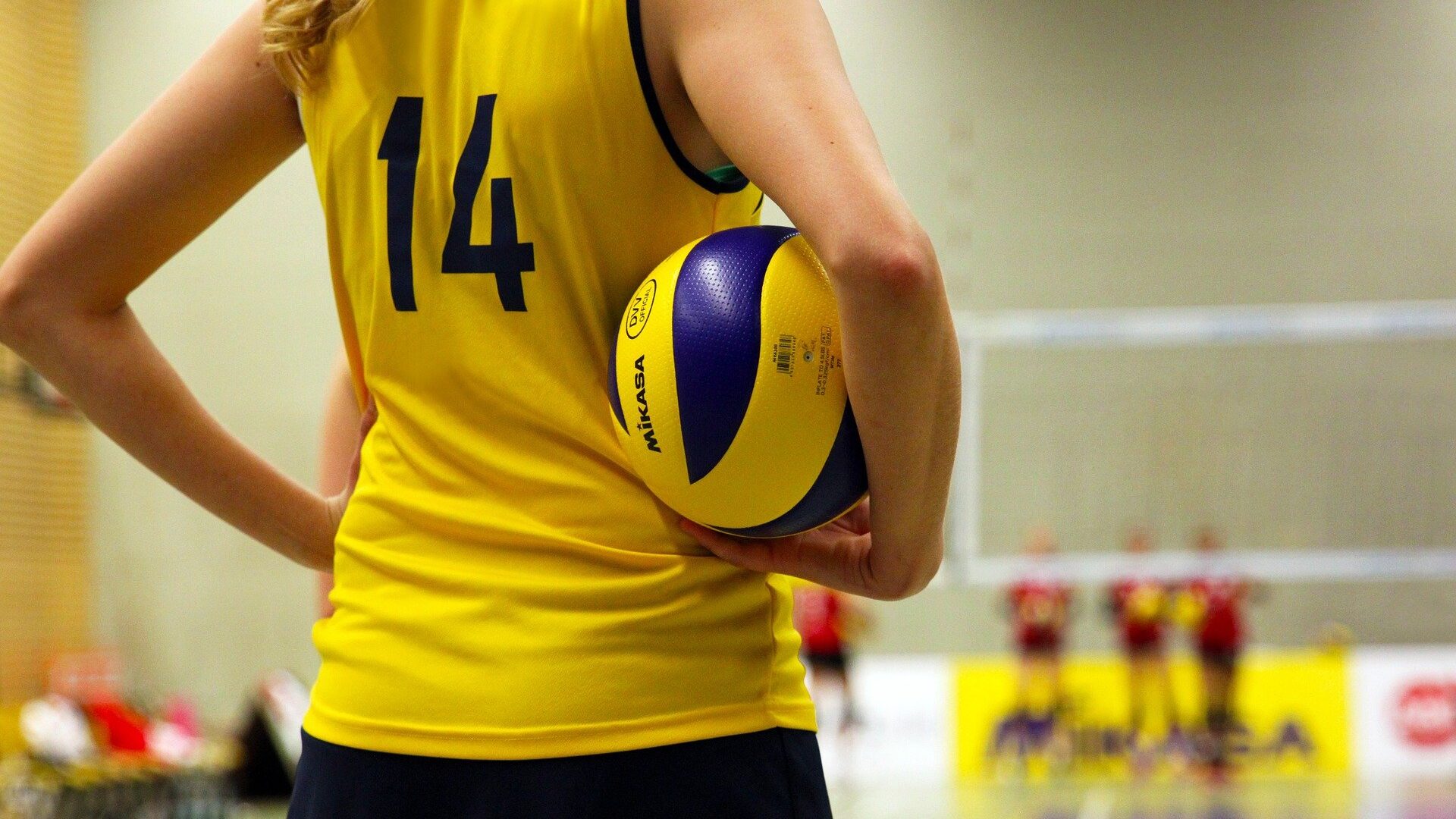 Abbigliamento sportivo di una ragazza che indossa una maglietta di pallavolo gialla con numero 14  tiene sotto il braccio una palla da pallavolo.