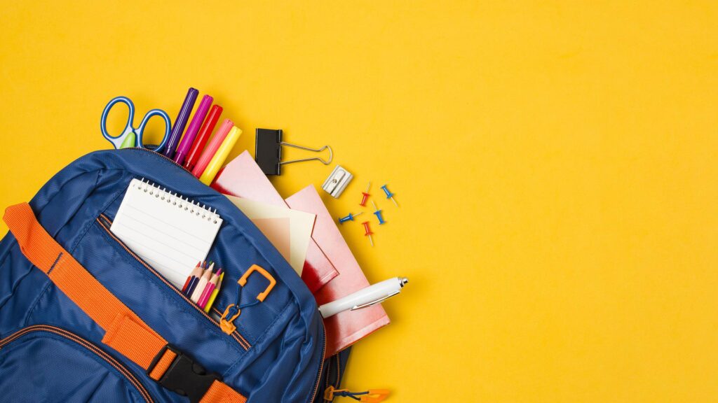 Materiale scolastico di cancelleria: zaino arancione e blu con dentro penne, matite, pennarelli, block notes, temperino, fogli, quaderno, puntine su tavolo giallo. 