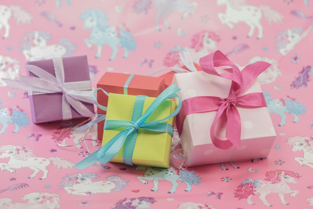 Materiale scolastico per la festa: 6 piccoli pacchi regalo di cartone colorato con nastri colorati su sfondo rosa.